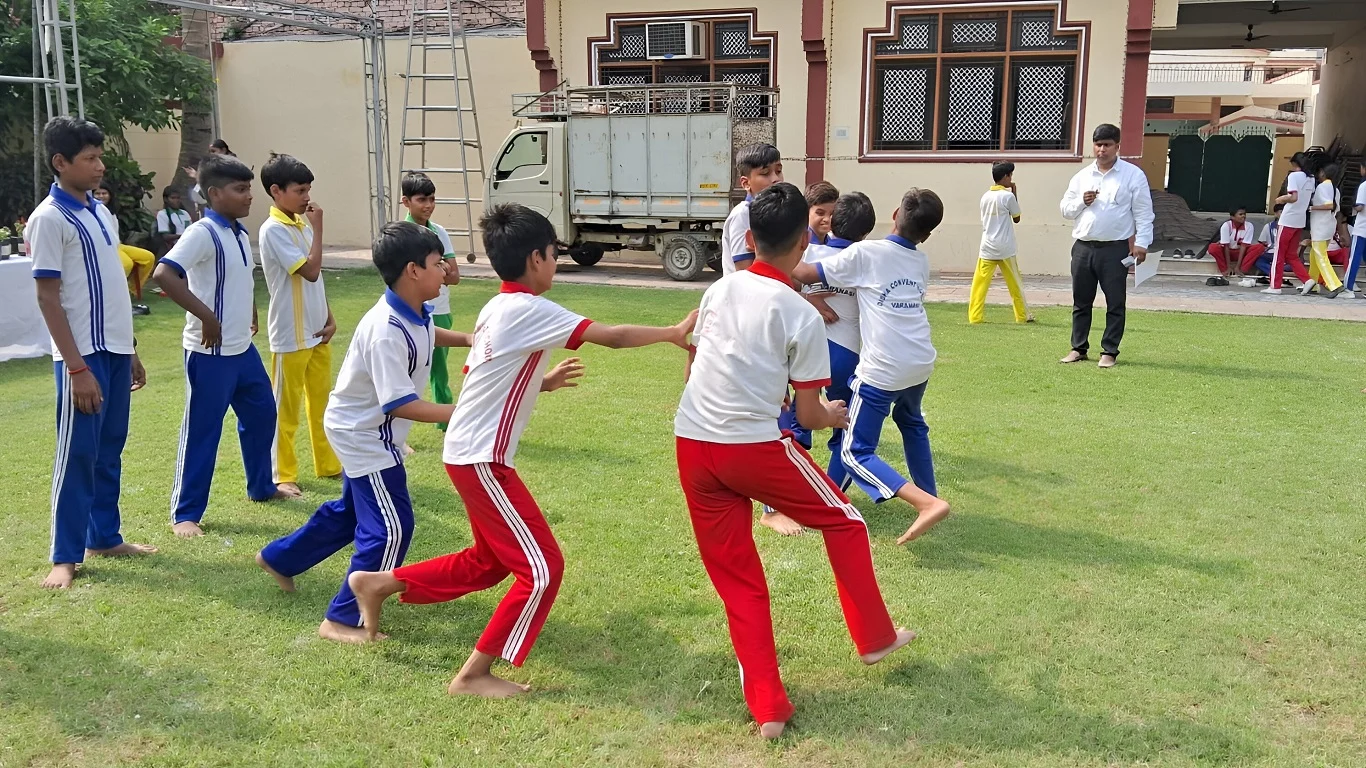 School children in sports uniforms playing a game on a grassy field, while a teacher observes in the background.